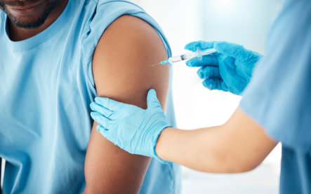 A healthcare worker in blue gloves gives a vaccine injection in a seated man’s upper arm, conveying a calm, routine medical visit.