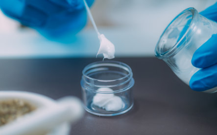 Blue-gloved hands use a thin applicator to place a white sample in a clear jar beside another container, suggesting careful laboratory testing.