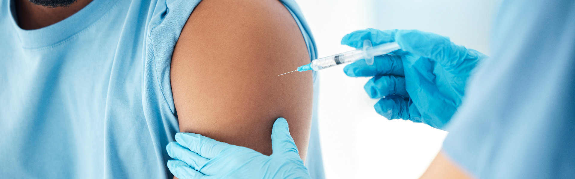 A healthcare worker in blue gloves gives a vaccine injection in a seated man’s upper arm, conveying a calm, routine medical visit.