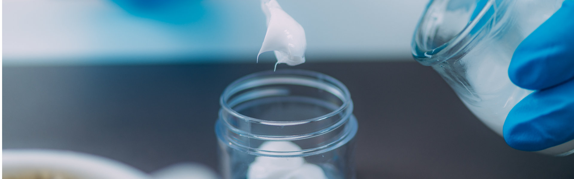 Blue-gloved hands use a thin applicator to place a white sample in a clear jar beside another container, suggesting careful laboratory testing.